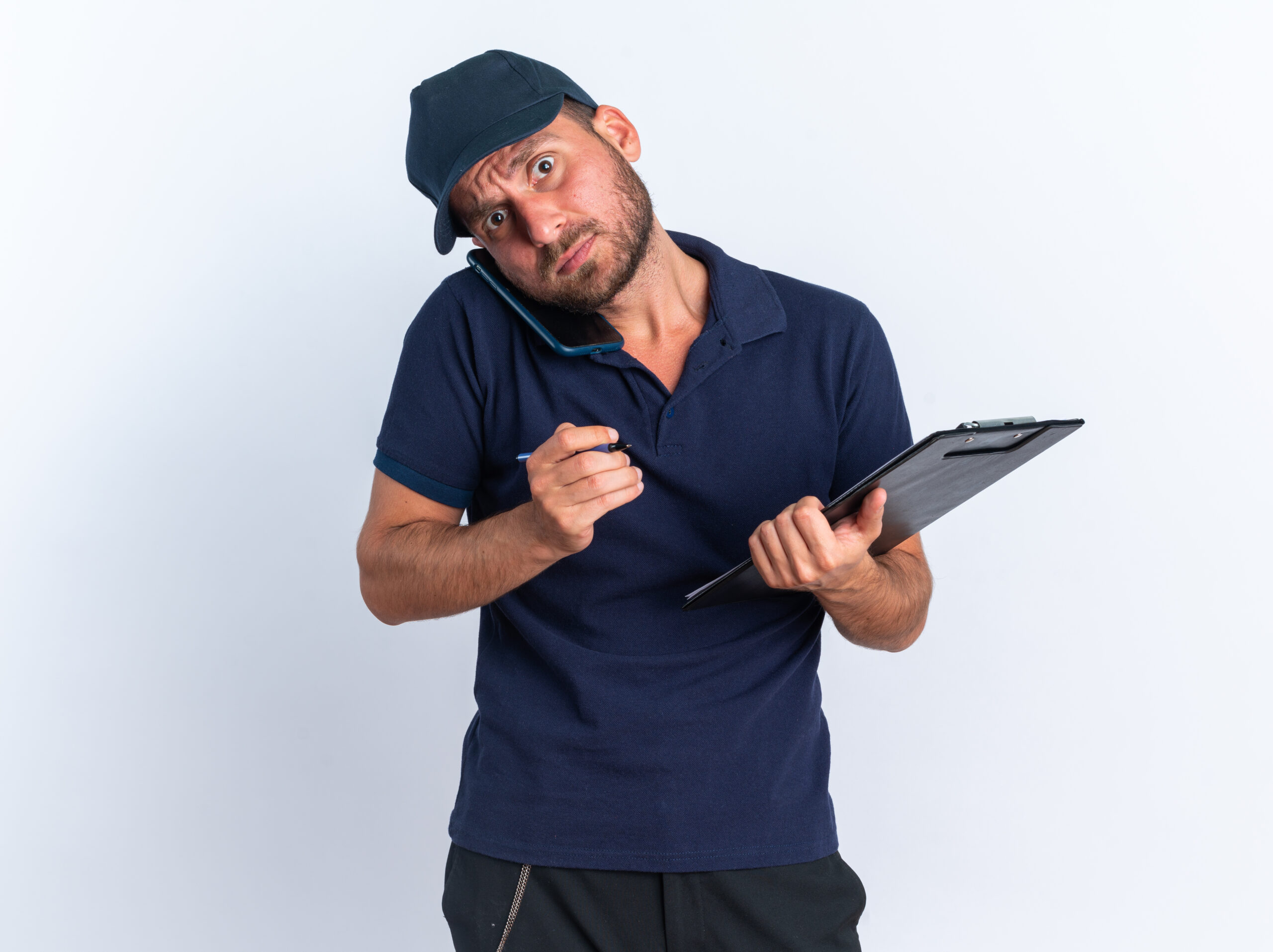 concerned young caucasian delivery man in blue uniform and cap holding pen and clipboard talking on phone looking at camera isolated on white background with copy space